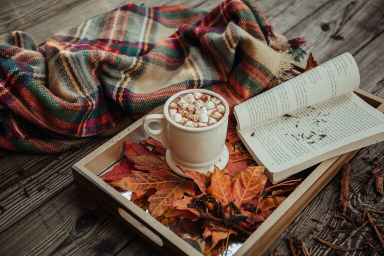 Cozy fall reading nook with a warm blanket, a cup of tea, and a book next to autumn leaves.